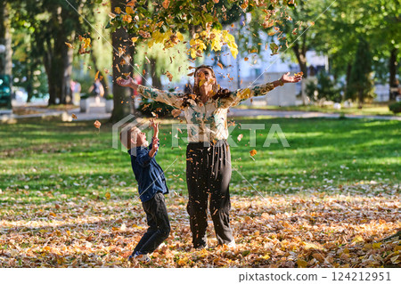 A modern woman joyfully plays with her son in the park, tossing leaves on a beautiful autumn day, capturing the essence of family life and the warmth of mother-son bonding in the midst of the fall 124212951