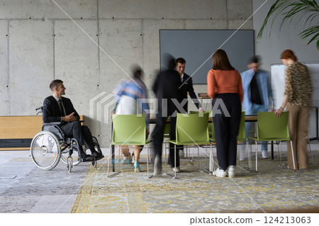 A diverse business team, working urgently around a conference table, collaborates on a meeting while their director, seated in a wheelchair, waits for their input 124213063