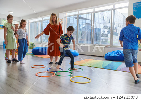 Small nursery school children with female teacher on floor indoors in classroom, doing exercise. Jumping over hula hoop circles track on the floor. 124213078