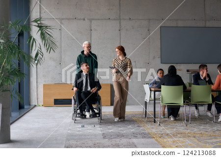 A diverse team of business colleagues, including a director in a wheelchair, engages in a collaborative discussion as they move together through a modern office space A diverse team of business colleagues, including a director in a wheelchair, engages in a collaborative discussion as they move together through a modern office space 124213088