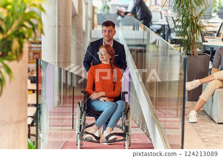 A company director assists his business colleague in a wheelchair, helping her navigate to their startup office, where they work alongside their diverse team of colleagues, emphasizing inclusivity and 124213089