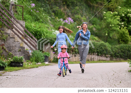 A cheerful little girl riding a balance bike with a helmet while two women joyfully run beside her on a paved path in a lush outdoor setting. 124213131