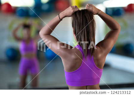 A determined woman ties her hair in preparation for an intense workout, embodying focus and strength as she gears up for her fitness training session A determined woman ties her hair in preparation for an intense workout, embodying focus and strength as she gears up for her fitness training session 124213312