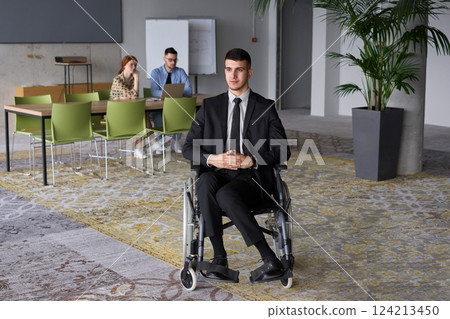 Business Director in a Suit Using a Wheelchair with Colleagues Conducting a Meeting Behind Him Business Director in a Suit Using a Wheelchair with Colleagues Conducting a Meeting Behind Him 124213450