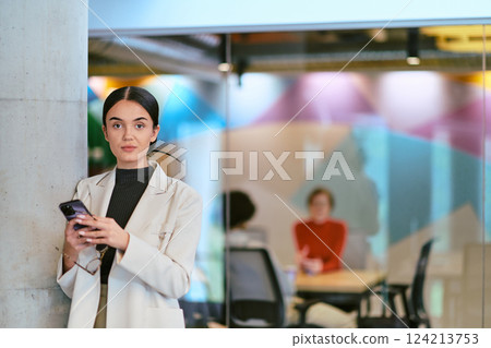In a modern glass office, a contemporary businesswoman effortlessly multitasks, using her smartphone, while surrounded by colleagues, illustrating the dynamics of corporate life and technology 124213753