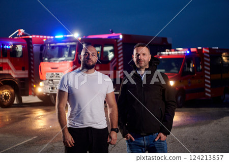 Group of firefighters, dressed in civilian clothing, stand in front of fire trucks during the night, showcasing a moment of camaraderie and unity among the team as they reflect on their duties and the Group of firefighters, dressed in civilian clothing, stand in front of fire trucks during the night, showcasing a moment of camaraderie and unity among the team as they reflect on their duties and the 124213857