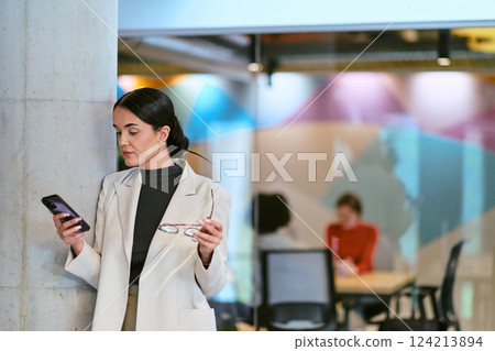 In a modern glass office, a contemporary businesswoman effortlessly multitasks, using her smartphone, while surrounded by colleagues, illustrating the dynamics of corporate life and technology In a modern glass office, a contemporary businesswoman effortlessly multitasks, using her smartphone, while surrounded by colleagues, illustrating the dynamics of corporate life and technology 124213894