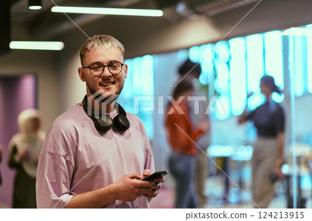 In a contemporary startup office, a modern blond entrepreneur stands, engrossed in his smartphone, epitomizing the dynamic and tech-savvy essence of the modern business world. 124213915