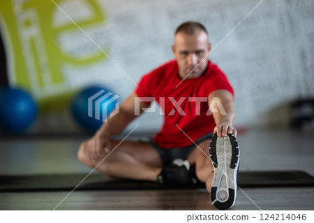 A fitness man stretches in the gym after an intense workout, focusing on recovery and flexibility. A fitness man stretches in the gym after an intense workout, focusing on recovery and flexibility. 124214046