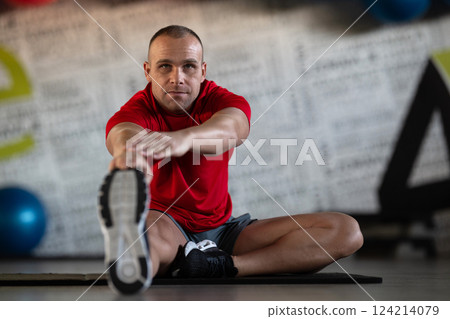 A fitness man stretches in the gym after an intense workout, focusing on recovery and flexibility. A fitness man stretches in the gym after an intense workout, focusing on recovery and flexibility. 124214079