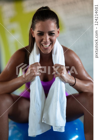 A beautiful and fit woman sitting on a gym ball with a towel, resting and recovering after an intense workout. A beautiful and fit woman sitting on a gym ball with a towel, resting and recovering after an intense workout. 124214081