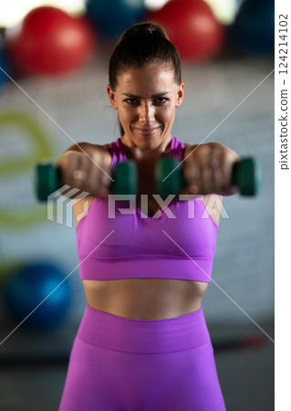 A focused and determined woman performs a strength building dumbbell workout at the gym, showcasing her athletic physique and dedication to fitness. 124214102