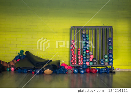 A neatly organized gym equipment storage area featuring a variety of weights, barbells, kettlebells, and fitness accessories against a yellow brick wall. A neatly organized gym equipment storage area featuring a variety of weights, barbells, kettlebells, and fitness accessories against a yellow brick wall. 124214111