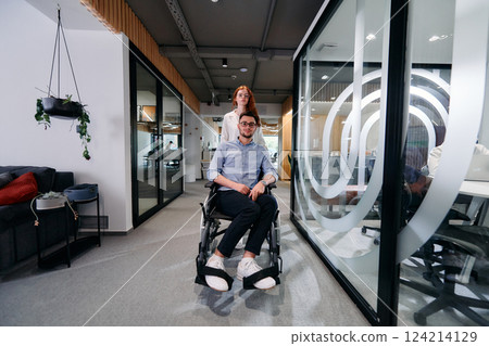 Business colleagues, collaborative business colleagues, including a person in a wheelchair, walk past a modern glass office corridor, illustrating diversity, teamwork and empowerment in the workplace. 124214129