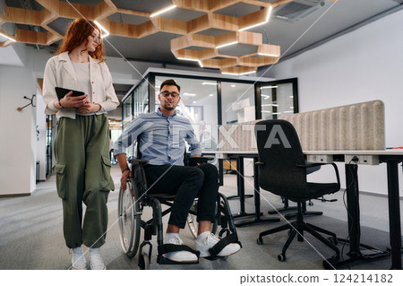Business colleagues, collaborative business colleagues, including a person in a wheelchair, walk past a modern glass office corridor, illustrating diversity, teamwork and empowerment in the workplace. 124214182
