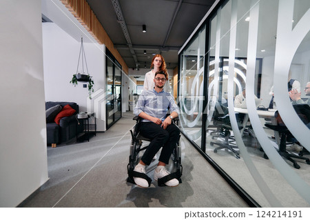 Business colleagues, collaborative business colleagues, including a person in a wheelchair, walk past a modern glass office corridor, illustrating diversity, teamwork and empowerment in the workplace. 124214191