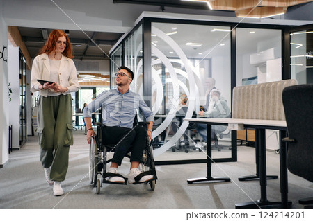 Business colleagues, collaborative business colleagues, including a person in a wheelchair, walk past a modern glass office corridor, illustrating diversity, teamwork and empowerment in the workplace. Business colleagues, collaborative business colleagues, including a person in a wheelchair, walk past a modern glass office corridor, illustrating diversity, teamwork and empowerment in the workplace. 124214201
