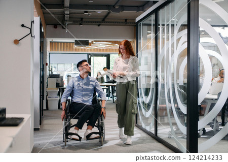 Business colleagues, collaborative business colleagues, including a person in a wheelchair, walk past a modern glass office corridor, illustrating diversity, teamwork and empowerment in the workplace. Business colleagues, collaborative business colleagues, including a person in a wheelchair, walk past a modern glass office corridor, illustrating diversity, teamwork and empowerment in the workplace. 124214233
