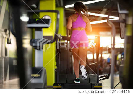 Woman Running on Treadmill in Gym.A fit woman wearing a purple sports outfit is running on a treadmill in a gym. She has a focused yet relaxed expression, with gym equipment and a warm light in the Woman Running on Treadmill in Gym.A fit woman wearing a purple sports outfit is running on a treadmill in a gym. She has a focused yet relaxed expression, with gym equipment and a warm light in the 124214446