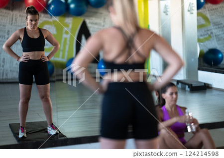 Two woman in the gym, one resting while the other stretches, recovering and recharging after an intense workout session. Two woman in the gym, one resting while the other stretches, recovering and recharging after an intense workout session. 124214598