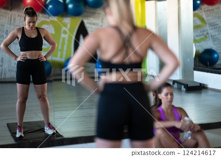 Two woman in the gym, one resting while the other stretches, recovering and recharging after an intense workout session. Two woman in the gym, one resting while the other stretches, recovering and recharging after an intense workout session. 124214617