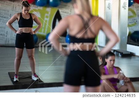 Two woman in the gym, one resting while the other stretches, recovering and recharging after an intense workout session. Two woman in the gym, one resting while the other stretches, recovering and recharging after an intense workout session. 124214635