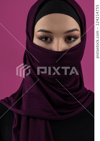 Beautiful Muslim woman in a hijab posing elegantly in a studio with a pink background. 124214755
