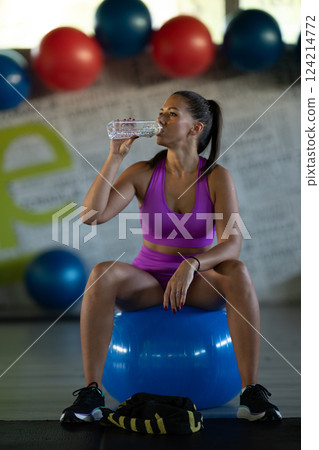 A fit woman takes a well deserved break in the gym, sipping water from her bottle after an intense workout session A fit woman takes a well deserved break in the gym, sipping water from her bottle after an intense workout session 124214772