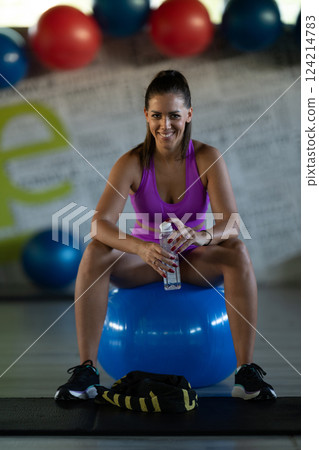 A fit woman takes a well deserved break in the gym, sipping water from her bottle after an intense workout session A fit woman takes a well deserved break in the gym, sipping water from her bottle after an intense workout session 124214783