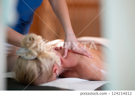 Relaxing Back Massage in a Spa Setting. A professional massage therapist in a blue uniform provides a relaxing back massage to a client lying on a massage table. 124214812