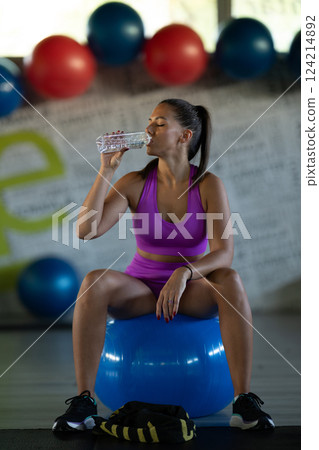 A fit woman takes a well deserved break in the gym, sipping water from her bottle after an intense workout session A fit woman takes a well deserved break in the gym, sipping water from her bottle after an intense workout session 124214892