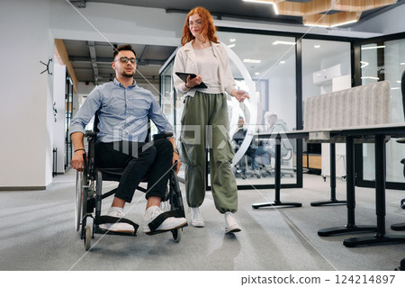 Business colleagues, collaborative business colleagues, including a person in a wheelchair, walk past a modern glass office corridor, illustrating diversity, teamwork and empowerment in the workplace. Business colleagues, collaborative business colleagues, including a person in a wheelchair, walk past a modern glass office corridor, illustrating diversity, teamwork and empowerment in the workplace. 124214897