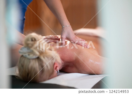 Relaxing Back Massage in a Spa Setting. A professional massage therapist in a blue uniform provides a relaxing back massage to a client lying on a massage table. 124214953