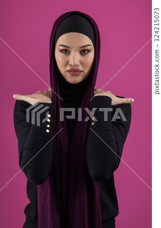 Beautiful Muslim woman in a hijab posing elegantly in a studio with a pink background. 124215073