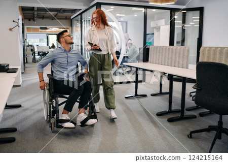 Business colleagues, collaborative business colleagues, including a person in a wheelchair, walk past a modern glass office corridor, illustrating diversity, teamwork and empowerment in the workplace. Business colleagues, collaborative business colleagues, including a person in a wheelchair, walk past a modern glass office corridor, illustrating diversity, teamwork and empowerment in the workplace. 124215164