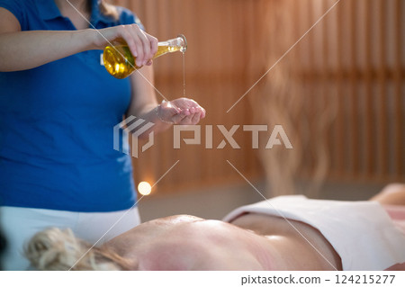 A professional massage therapist pours aromatic oil into their hands, preparing for a relaxing and rejuvenating massage session in a serene spa setting. A professional massage therapist pours aromatic oil into their hands, preparing for a relaxing and rejuvenating massage session in a serene spa setting. 124215277