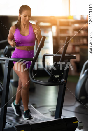 Woman Running on Treadmill in Gym.A fit woman wearing a purple sports outfit is running on a treadmill in a gym. She has a focused yet relaxed expression, with gym equipment and a warm light in the 124215316