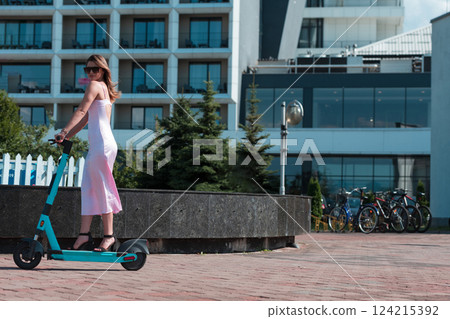 Elegant woman in a pink dress and sunglasses riding an electric scooter through the sunny city streets, embracing the freedom and style of a perfect urban adventure. Elegant woman in a pink dress and sunglasses riding an electric scooter through the sunny city streets, embracing the freedom and style of a perfect urban adventure. 124215392