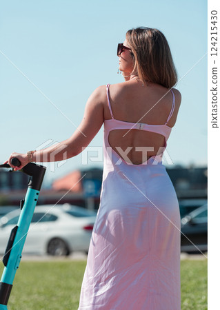 An elegant, stunning woman in a pink dress and sunglasses strikes a confident pose next to her electric scooter, radiating style and sophistication in the vibrant urban environment. An elegant, stunning woman in a pink dress and sunglasses strikes a confident pose next to her electric scooter, radiating style and sophistication in the vibrant urban environment. 124215430