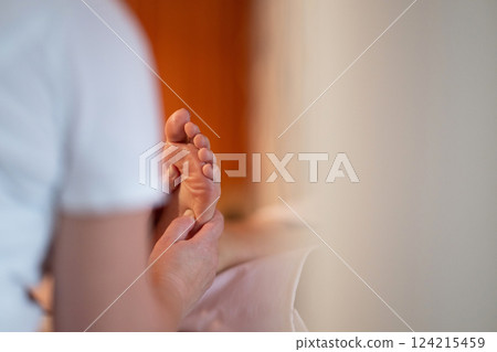 Foot Massage Therapy in a Relaxing Spa Setting. A professional massage therapist in a white uniform provides a soothing foot massage to a client in a calm and serene spa environment 124215459