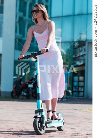 Elegant woman in a pink dress and sunglasses riding an electric scooter through the sunny city streets, embracing the freedom and style of a perfect urban adventure. 124215518