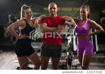 A determined fitness trainer standing confidently in front of his two female clients, guiding them through their workout in the background. A determined fitness trainer standing confidently in front of his two female clients, guiding them through their workout in the background. 124215582