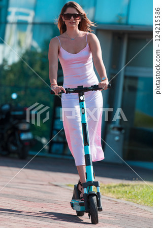 Elegant woman in a pink dress and sunglasses riding an electric scooter through the sunny city streets, embracing the freedom and style of a perfect urban adventure. Elegant woman in a pink dress and sunglasses riding an electric scooter through the sunny city streets, embracing the freedom and style of a perfect urban adventure. 124215586