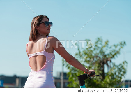 An elegant, stunning woman in a pink dress and sunglasses strikes a confident pose next to her electric scooter, radiating style and sophistication in the vibrant urban environment. An elegant, stunning woman in a pink dress and sunglasses strikes a confident pose next to her electric scooter, radiating style and sophistication in the vibrant urban environment. 124215602