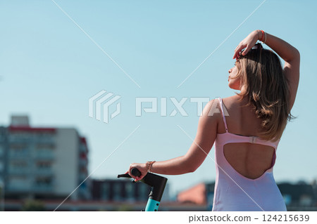 An elegant, stunning woman in a pink dress and sunglasses strikes a confident pose next to her electric scooter, radiating style and sophistication in the vibrant urban environment. An elegant, stunning woman in a pink dress and sunglasses strikes a confident pose next to her electric scooter, radiating style and sophistication in the vibrant urban environment. 124215639