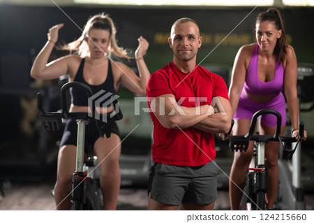 A determined fitness trainer standing confidently in front of his two female clients, guiding them through their workout in the background. A determined fitness trainer standing confidently in front of his two female clients, guiding them through their workout in the background. 124215660