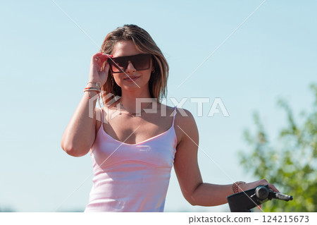 An elegant, stunning woman in a pink dress and sunglasses strikes a confident pose next to her electric scooter, radiating style and sophistication in the vibrant urban environment. 124215673
