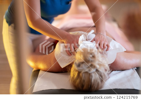 Relaxing Back Massage in a Spa Setting. A professional massage therapist in a blue uniform provides a relaxing back massage to a client lying on a massage table. 124215769