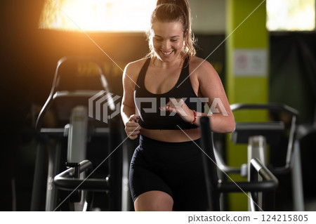 Woman Running on Treadmill in Gym.A fit woman wearing a purple sports outfit is running on a treadmill in a gym. She has a focused yet relaxed expression, with gym equipment and a warm light in the 124215805