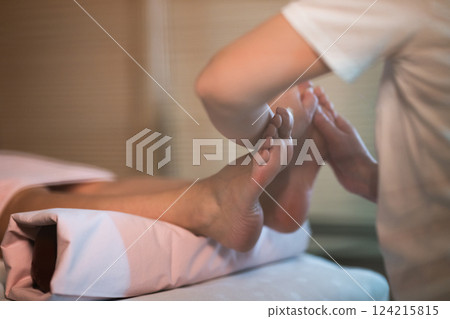 Foot Massage Therapy in a Relaxing Spa Setting. A professional massage therapist in a white uniform provides a soothing foot massage to a client in a calm and serene spa environment Foot Massage Therapy in a Relaxing Spa Setting. A professional massage therapist in a white uniform provides a soothing foot massage to a client in a calm and serene spa environment 124215815